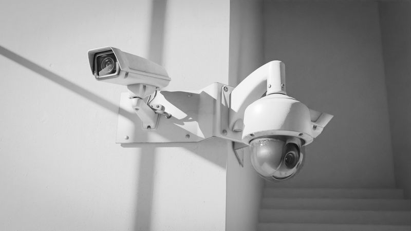 Two security cameras mounted on a white wall, one is a dome camera and the other a box camera, casting shadows in bright light.