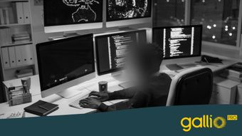 Person, die an einem Schreibtisch mit mehreren Computerbildschirmen arbeitet, auf denen Code angezeigt wird, in einem schwach beleuchteten Büro mit Regalen und großen Fenstern.