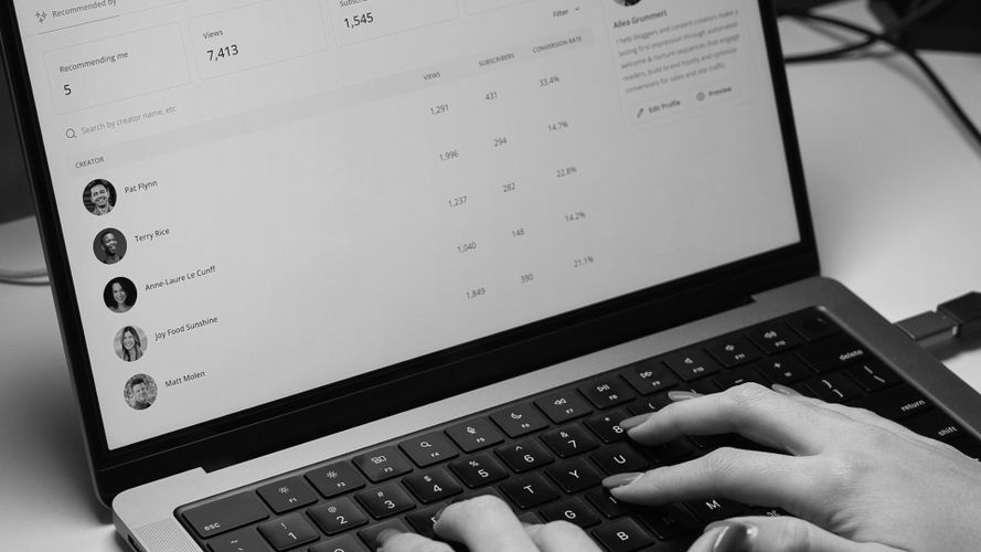 Dłonie piszące na klawiaturze laptopa, na którego ekranie widoczny jest czarno-biały pulpit nawigacyjny z danymi użytkowników i analizami.
