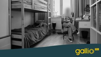 Una habitación de dormitorio moderna y monocromática con literas, un escritorio y una ventana. Una persona está sentada, borrosa por privacidad, en una silla junto al escritorio.