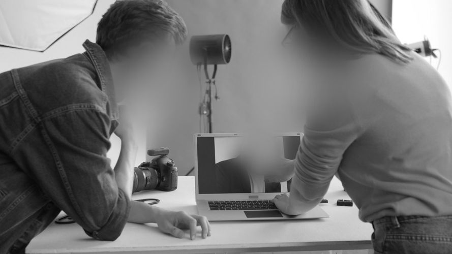 Dwie osoby w studiu przeglądają zdjęcia na laptopie. Widać aparat fotograficzny i sprzęt oświetleniowy. Twarze są zamazane.