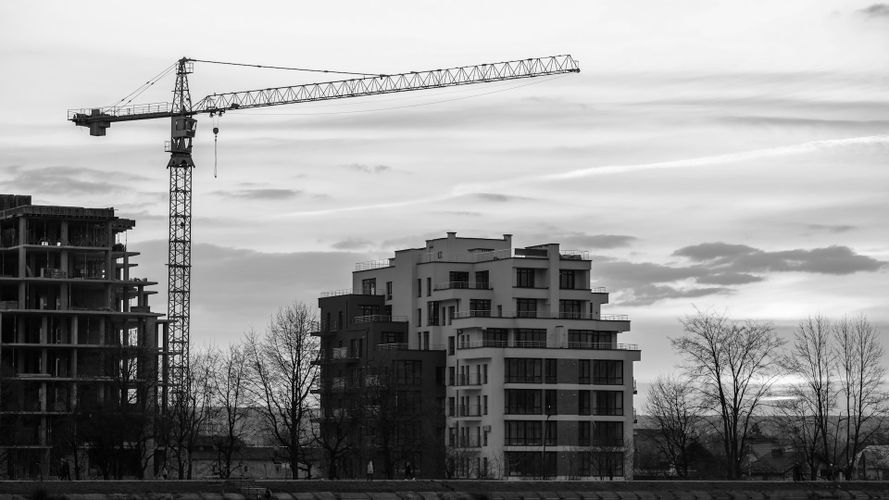 Czarno-białe zdjęcie żurawia obok dwóch nowoczesnych budynków pod zachmurzonym niebem, z gołymi drzewami na pierwszym planie.