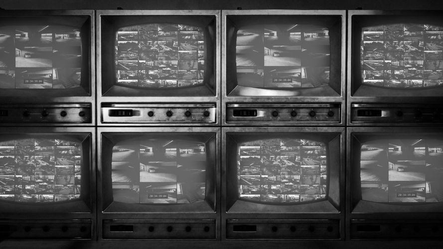 Czarno-biały obraz ośmiu zabytkowych telewizorów CRT pokazujących nagrania z monitoringu, ułożonych w siatkę dwa na cztery.