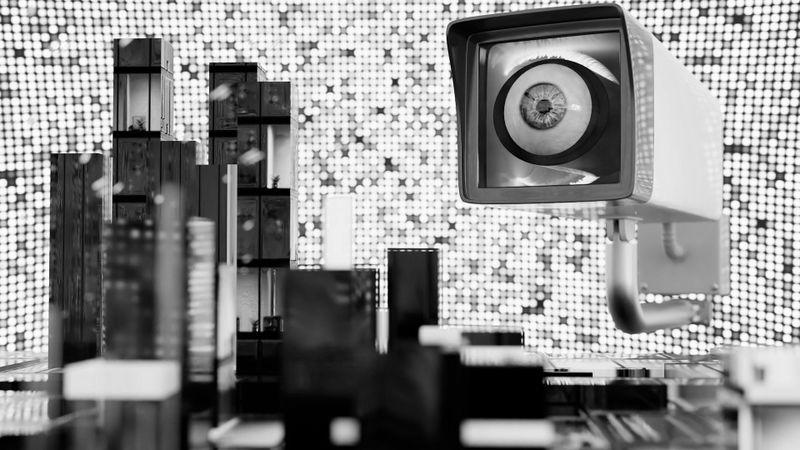Escena surrealista en blanco y negro con una gran cámara de vigilancia que supervisa modelos abstractos de paisajes urbanos contra un fondo pixelado.