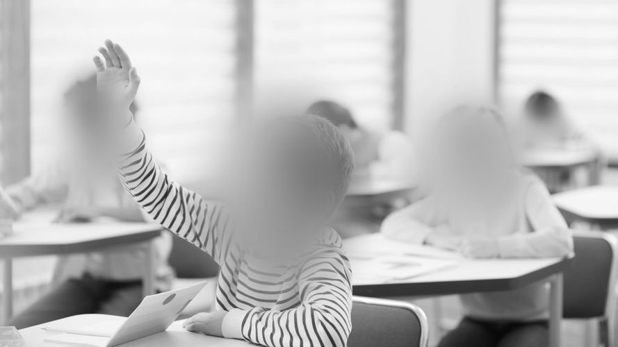 Uczeń w koszuli w paski podnosi rękę w klasie, podczas gdy inni pracują przy ławkach. Twarze są zamazane ze względu na ochronę prywatności.