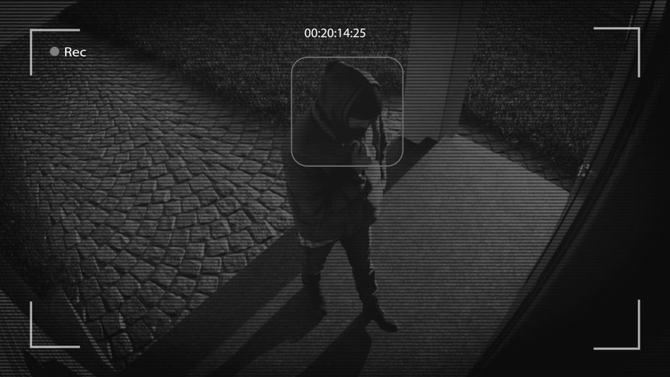 Security camera footage of a person in a hooded jacket standing outside on a cobblestone path, viewed through a black-and-white filter.