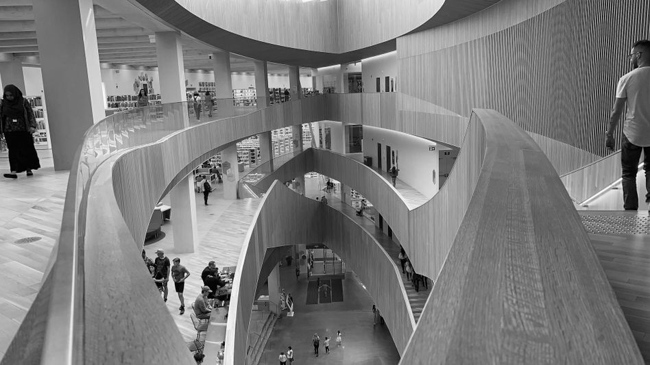 Image en noir et blanc d'un intérieur de bibliothèque moderne à plusieurs niveaux avec des passerelles en bois courbes et des personnes marchant sur différents étages.