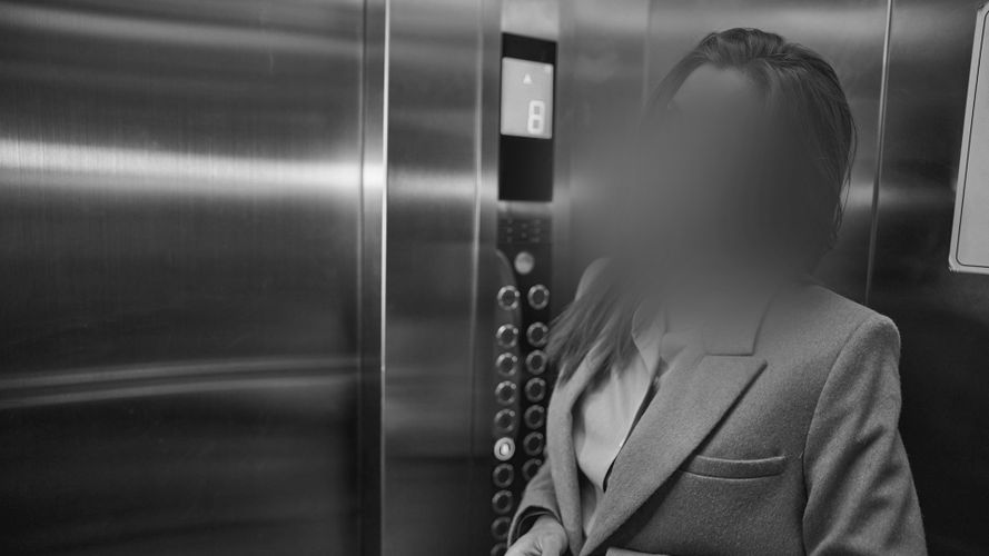 Osoba w garniturze stojąca w windzie, z zamazaną twarzą; z boku widoczne są przyciski i panel windy.