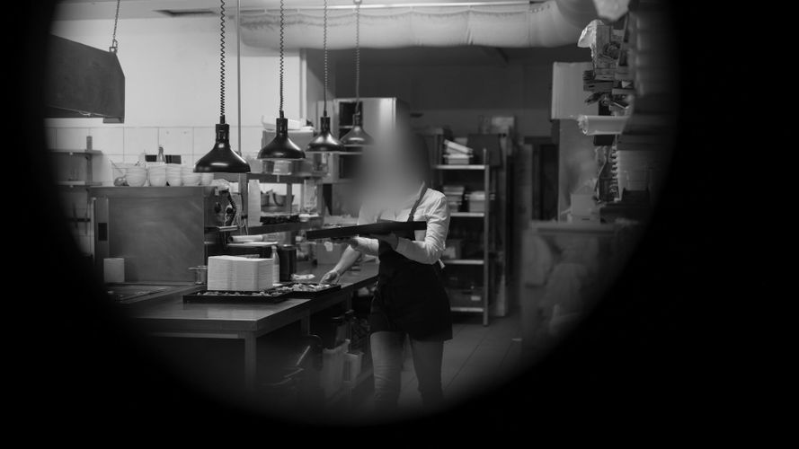 Persona con delantal llevando una bandeja en una cocina profesional, rodeada de equipo industrial y luces colgantes. La imagen tiene viñetas.