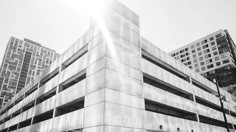 Photo en noir et blanc d’un parking en béton à plusieurs niveaux, avec des tours résidentielles en arrière-plan.
