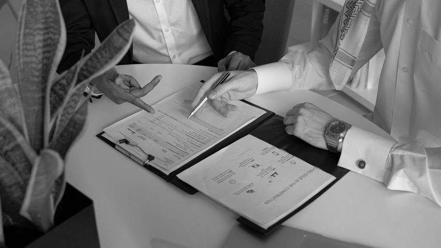 Dos personas discutiendo documentos sobre una mesa, con una señalando y la otra sosteniendo un bolígrafo, junto a una planta. Parecen estar vestidas de manera profesional.
