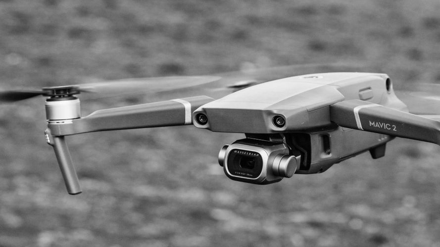 Zbliżenie na dron DJI Mavic 2 w locie, pokazujące jego kamerę i śmigła, na tle rozmytego krajobrazu na zewnątrz. Obraz w czerni i bieli.