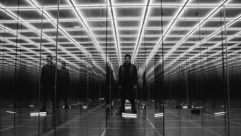 Person standing in a mirrored room with a grid of bright lights on the ceiling, creating multiple reflections.
