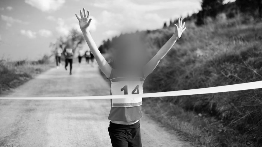 Dziecko w koszulce z numerem 14 przekracza linię mety z uniesionymi rękami na wiejskiej ścieżce podczas biegu, a w tle widać zamazane sylwetki innych biegaczy. Zdjęcie czarno-białe.