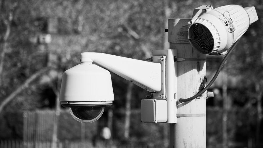 Czarno-białe zdjęcie kamery bezpieczeństwa zamontowanej na słupie, z zamazanym tłem drzew i ogrodzenia.