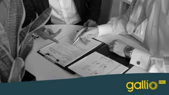 Dos personas con ropa formal discuten documentos sobre una mesa, señalando papeles en una carpeta, con una planta cerca.