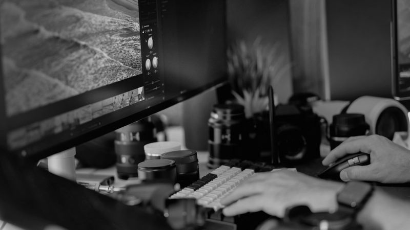 Fotografía en escala de grises de un escritorio: manos sobre un teclado y un ratón, cámara y objetivos junto a un monitor en el que se muestra un programa de edición fotográfica.