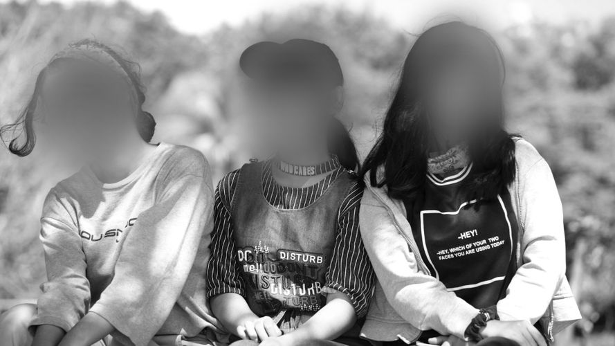 Trzy osoby siedzące obok siebie na zewnątrz, twarze rozmazane, ubrane w swobodne ubrania. W tle rozmazana zieleń. Czarno-biały obraz.