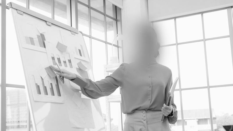La foto en blanco y negro fue sometida a un proceso de anonimización. Muestra a una mujer señalando gráficos en una pizarra