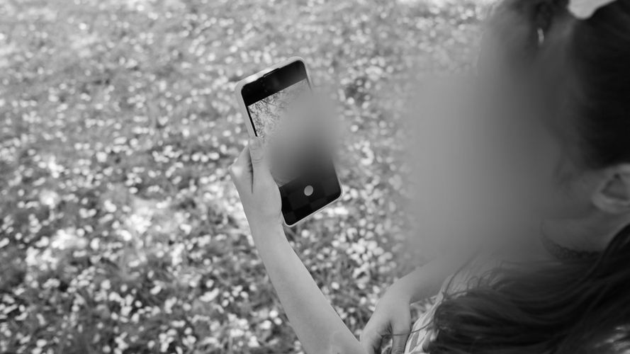 Osoba trzymająca smartfon na zewnątrz, prawdopodobnie robiąca zdjęcie trawie i kwiatom. Twarz tej osoby jest rozmyta.