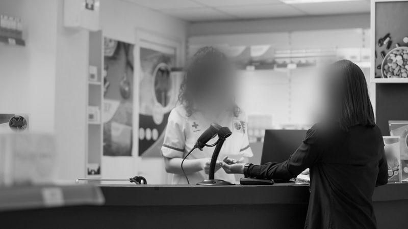 Two people at a pharmacy counter, one handing a card to the other. Faces are blurred. Shelves with products are visible in the background.
