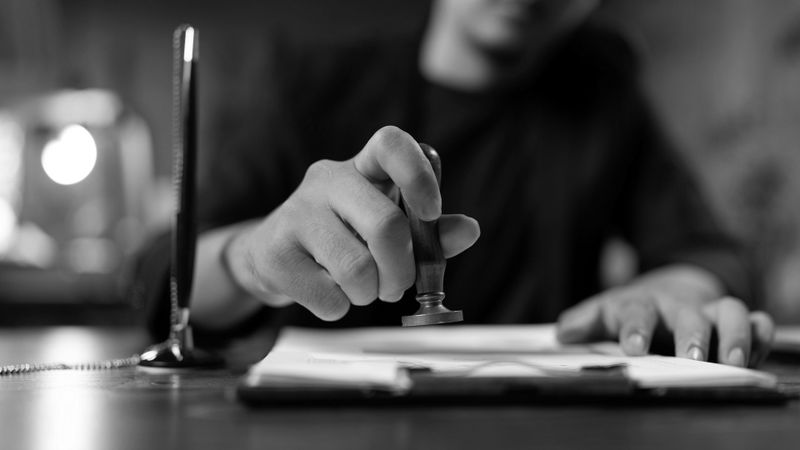 Une personne tamponne un document sur un bureau, tenant un tampon d'une main et reposant l'autre sur le papier. Photo en noir et blanc.