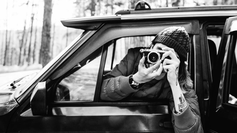 foto en blanco y negro de un hombre con tatuajes asomándose por la ventana del lado del conductor y tomando una foto con una cámara