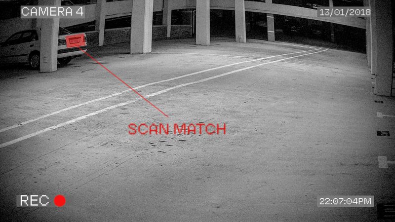 Black and white surveillance footage showing a parking garage with a car in the distance. Text "SCAN MATCH" is highlighted near the car.