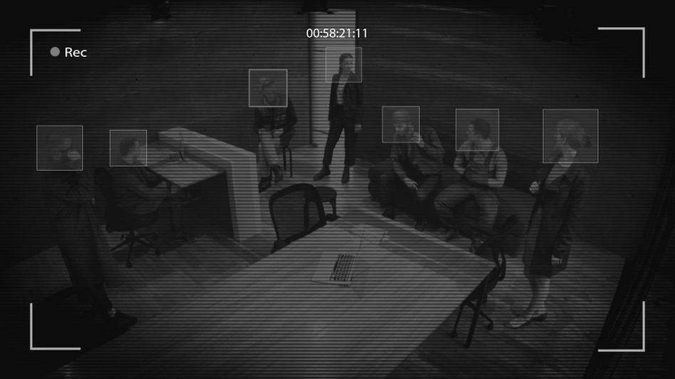 Surveillance footage of a group meeting in an office, with several people sitting and standing around a table.