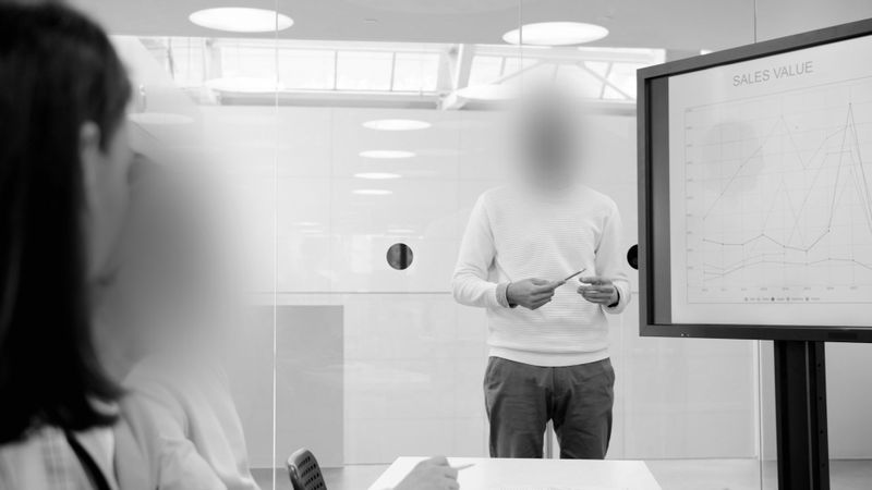 Dos empleadas están sentadas en una mesa con una pantalla donde se muestran los resultados de ventas, un empleado que dirige la reunión las observa y les habla, foto en blanco y negro