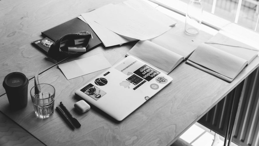 Una imagen en blanco y negro de un escritorio de madera con una laptop, papeles dispersos, un bolígrafo, una taza de café, un vaso de agua y una bolsa para laptop.