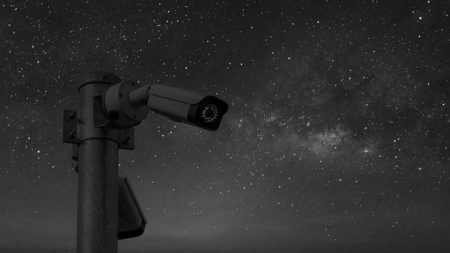 Zamontowana na słupie kamera monitoringu widoczna jest jako sylwetka na tle rozgwieżdżonego nocnego nieba, w którym widać Drogę Mleczną.