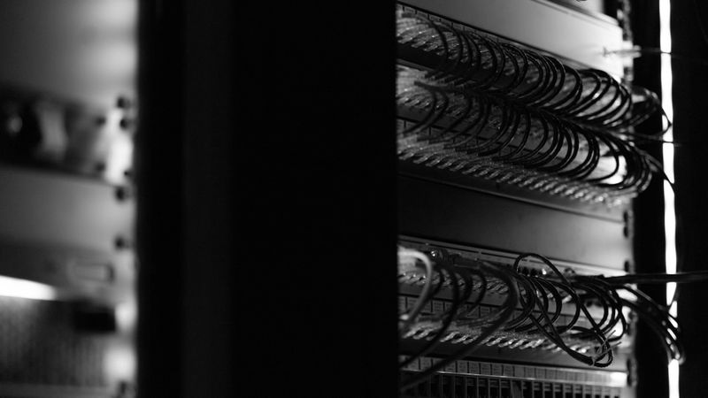 Foto en blanco y negro de un rack de servidores con los cables organizados cuidadosamente, iluminado por una luz mínima, creando una atmósfera de alto contraste y con un toque melancólico.