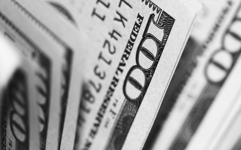 Gros plan de plusieurs billets de cent dollars américains superposés en noir et blanc, montrant les détails des chiffres et du texte imprimés.