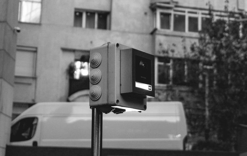foto en blanco y negro de la ciudad, en el centro una cámara de vigilancia doméstica y detrás un furgón blanco y un bloque de apartamentos