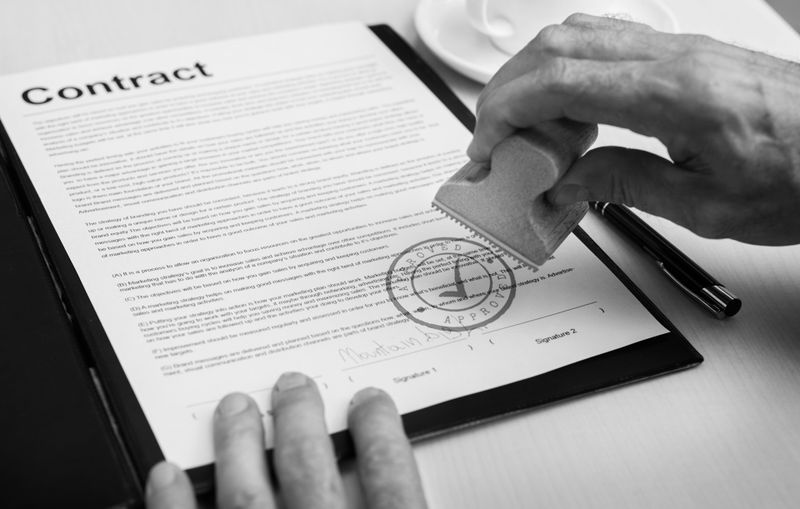 Image en noir et blanc d'une personne apposant un sceau officiel sur un document contractuel, avec un stylo et une tasse de thé à proximité.