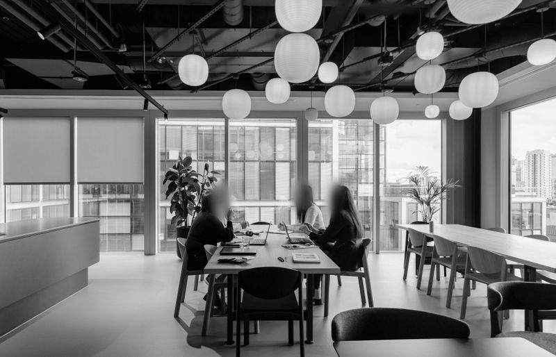 Black and white office scene with three blurred figures at a table, large windows, and round ceiling lights. Cityscape visible outside.