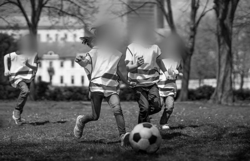 Children playing soccer on a grassy field, wearing striped jerseys. The scene is in black and white, with blurred faces and trees in the background.