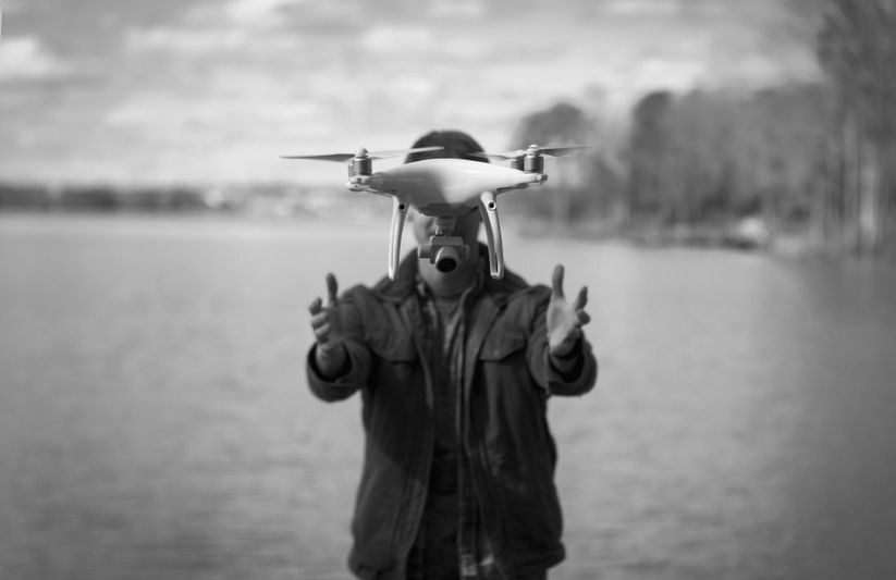 Persona junto a un lago, acercándose a un dron flotante, con árboles y cielo nublado al fondo. Imagen en blanco y negro.