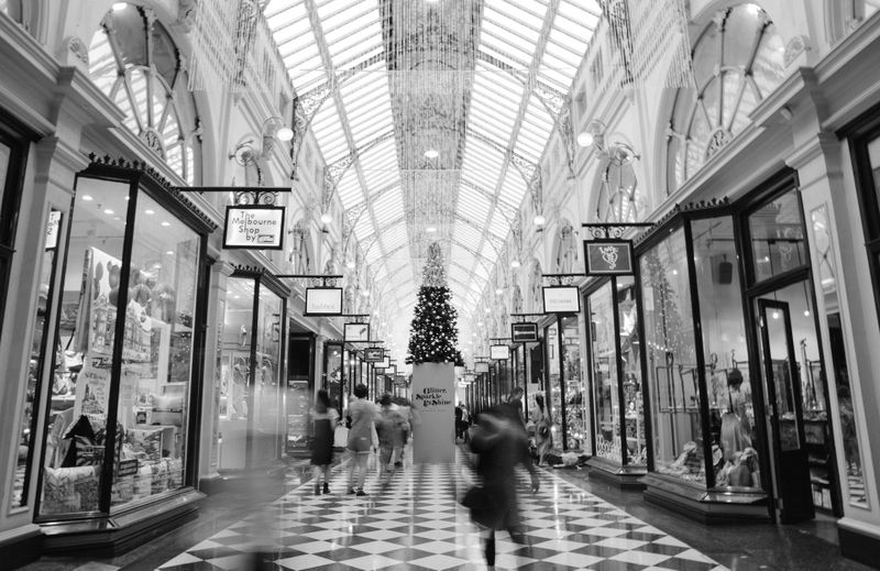 Black and white image of a bustling shopping mall with high arched ceilings, a central Christmas tree, and shoppers walking along a checkered floor.