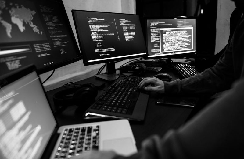 Person, die an einem Schreibtisch mit mehreren Computermonitoren sitzt, auf denen in einem schwach beleuchteten Raum Codezeilen angezeigt werden.