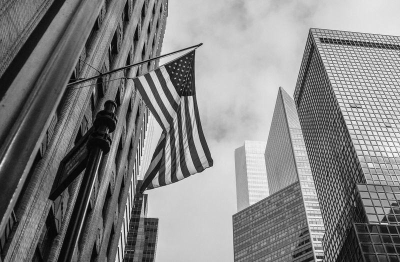 Black and white photo of American flag on a pole against a backdrop of tall skyscrapers under a cloudy sky.