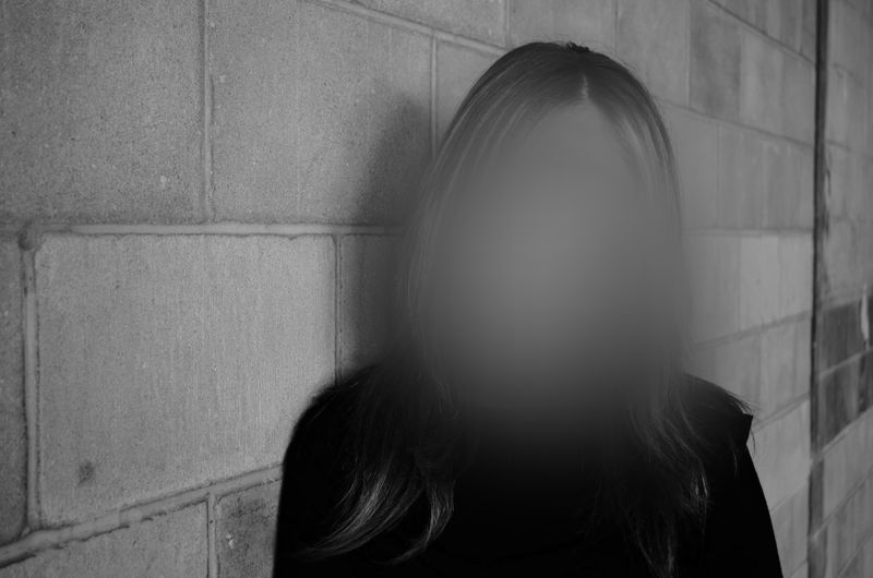 A person with a blurred face stands against a concrete block wall, wearing a dark top. The image is in black and white.