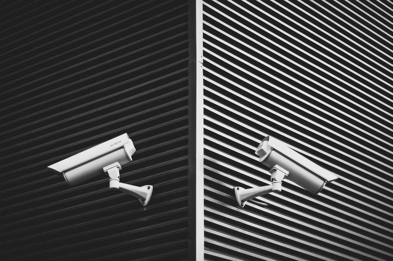 Deux caméras de sécurité montées sur un mur rayé et angulaire, orientées dans des directions opposées, dans une image en noir et blanc.