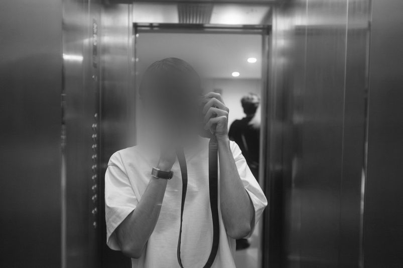 Une personne prend un selfie dans un ascenseur, le visage flou, vêtue d'une chemise blanche et d'une montre. Une autre personne est visible en arrière-plan. Noir et blanc.