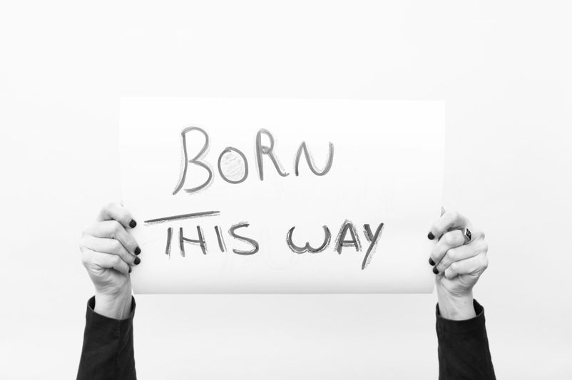 Des mains avec du vernis à ongles noir tiennent une pancarte sur laquelle on peut lire « BORN THIS WAY » sur un fond blanc uni.