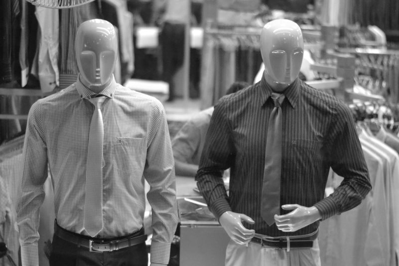 Deux mannequins en chemise et cravate exposés dans un magasin de vêtements, entourés de portants. Image en noir et blanc.