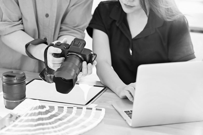 Deux personnes examinent un appareil photo tandis que l'une utilise un ordinateur portable, avec un objectif et des échantillons de couleurs sur la table. Image en noir et blanc.