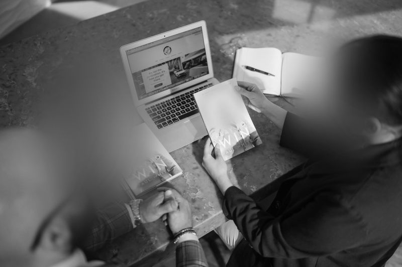 Two people discuss while looking at a laptop and booklet, with a notebook open on a table. The image is in black and white.