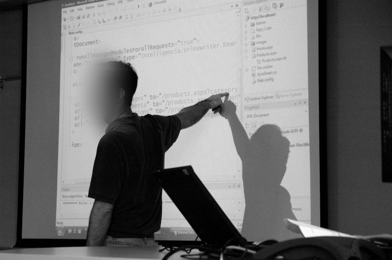 Person, die während einer Präsentation auf Code auf einer Projektionsfläche zeigt, wobei im Vordergrund ein Laptop sichtbar ist.